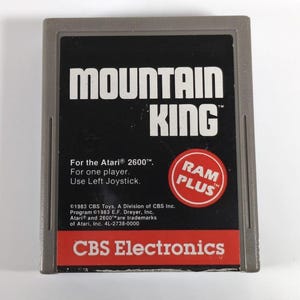 May include: A vintage Mountain King video game cartridge for the Atari 2600. The cartridge has a gray frame with a black label featuring the game title in white block letters. The label also includes the text "RAM PLUS" in a red circle and "CBS Electronics".
