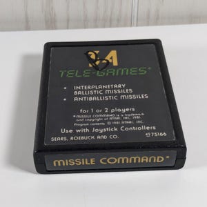 May include: Black Tele-Games Missile Command video game cartridge. The label includes the game title, "Missile Command," and details about the game's features, such as interplanetary ballistic missiles. The cartridge is designed for use with joystick controllers.