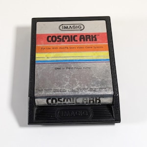 May include: A black video game cartridge for the Atari and Sears systems. The label features the title "COSMIC ARK" in red, with orange, yellow, blue, and purple stripes. The cartridge is labeled "ONE OR TWO Player Game."