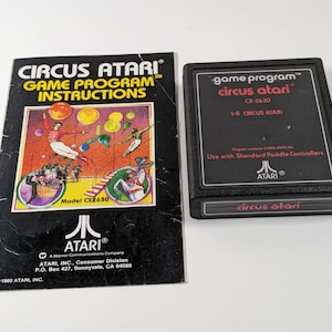 May include: An Atari Circus Atari game cartridge and instruction manual. The black cartridge has the game title and CX-2630 model number. The manual features colorful circus-themed graphics and the Atari logo.