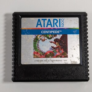May include: A black Atari 5200 game cartridge with a silver label. The label features the Atari logo, the game title "CENTIPEDE", and a colorful illustration of the game's characters. The copyright information reads "© 1982 ATARI, INC. ALL RIGHTS RESERVED 5215".