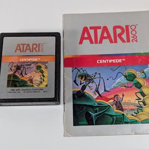 May include: An Atari 2600 Centipede video game cartridge with a black plastic casing and silver label, alongside its matching instruction manual. Both items feature the red Atari 2600 logo and the Centipede game title with colorful artwork.