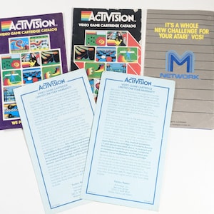 May include: Four brochures for Activision video game cartridges. The brochures are blue, purple, and gray with black text. The text on the brochures includes "Activision", "Video Game Cartridge Catalog", "Limited One Year Warranty", and "It's a whole new challenge for your Atari VCS!"