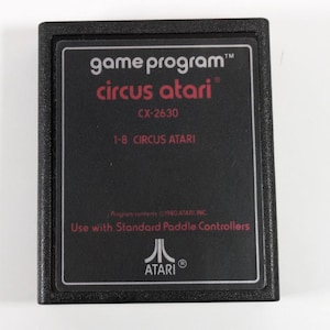 May include: Black Atari game cartridge with the text "game program" "circus atari" "CX-2630" "1-8 CIRCUS ATARI" "TM" "Program contents 1980 ATARI, INC." "Use with Standard Paddle Controllers" "ATARI" "®" printed on it.