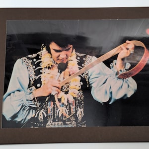 May include: A close-up photo of Elvis Presley wearing a blue shirt with a beaded vest and a lei. He is holding a microphone and a brown leather strap.