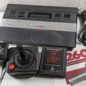 May include: A black Atari 2600 video game console with a joystick and a game cartridge labeled "game program combat CX-2601". The cartridge lists the different game options, including "Tank", "Tank Pong", "The Invisible Tank", "Plane", and "Jet Fighters".