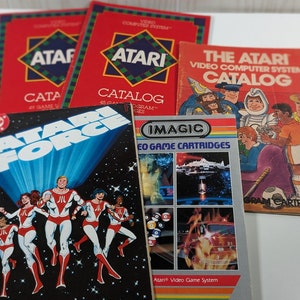 May include: A collection of vintage Atari video game catalogs and a cartridge case. The catalogs feature the Atari logo and the text "The Atari Video Computer System Catalog". The cartridge case features a colorful illustration of a spaceship and the text "Imagic Video Game Cartridges".