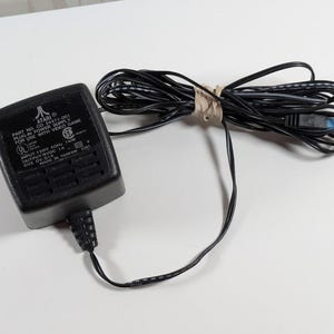 May include: A black Atari power supply adapter with a long, coiled black cord secured by a tan rubber band. The rectangular block features white printed text including Part No. CO 24471-001, Input 120V 60Hz 15W, and Output 9VDC 1A, with a blue-tipped connector.