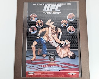 May include: Framed UFC Ultimate Fighting Championship action figure display. The image features two action figures in a wrestling hold, with the UFC logo and text "AS REAL AS IT GETS." The display is in a brown frame.