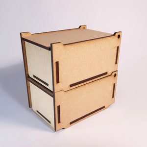 Könnte beinhalten: Zwei übereinander gestapelte Holzkisten mit hellbrauner Oberfläche und dunkelbraunen Verzierungen. Die Kisten haben ein schlichtes Design und eignen sich perfekt zum Organisieren kleiner Gegenstände.