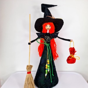 May include: A felt witch doll with long red hair, wearing a black dress with a spiderweb design, a black hat with a feather, and holding a red bag and a broom.