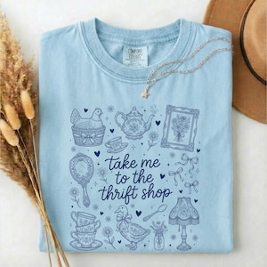 May include: Light blue t-shirt with a whimsical design featuring a hen in a basket, teacups, a teapot, and the phrase "take me to the thrift shop" in a handwritten font. A star pendant necklace and a brown hat are also visible.