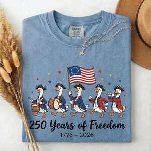 May include: A blue jean colored t-shirt featuring a marching band of geese in colonial attire, carrying a flag. The shirt has the text "250 Years of Freedom 1776-2026". A gold necklace and a brown hat are in the background.