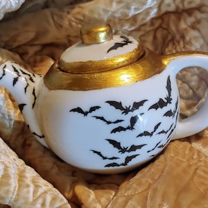 May include: A white ceramic teapot with a gold lid and black bat silhouettes. The teapot has a rounded body and a small spout.