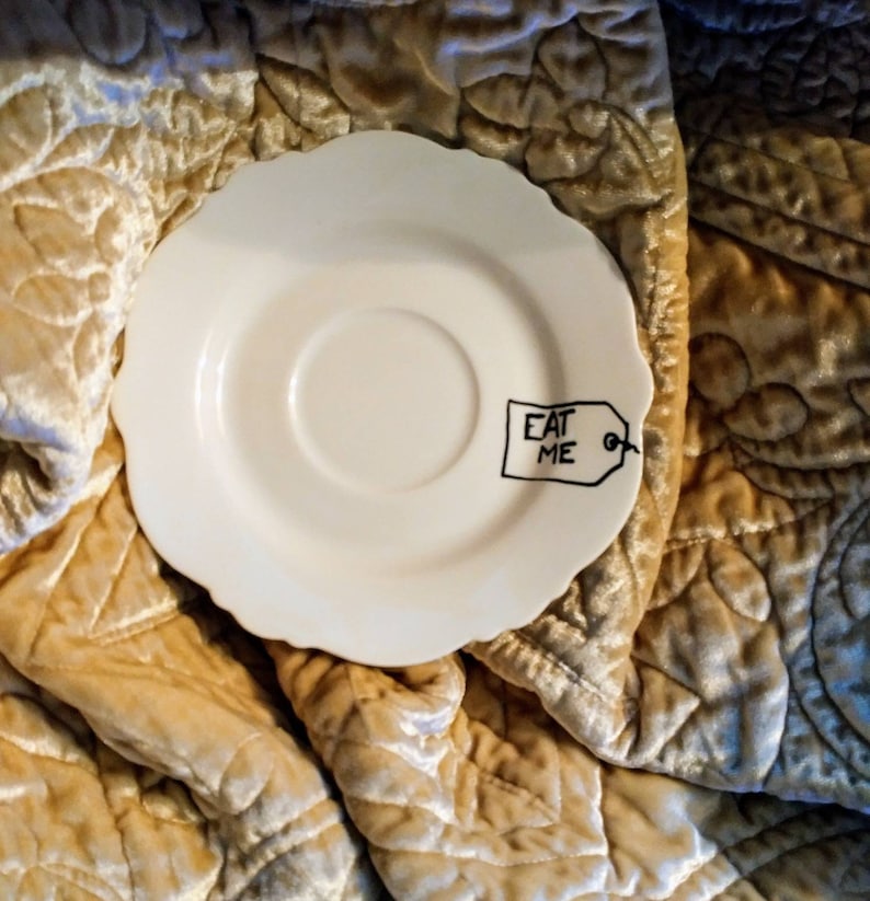 May include: White ceramic saucer with a black tag that says "Eat Me" on it. The saucer is on a gold quilted fabric.