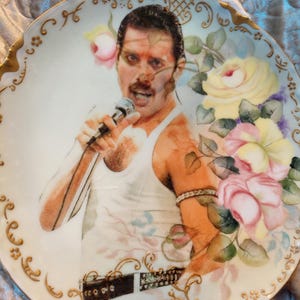 May include: A decorative porcelain plate with a portrait of a singer in a white vest, holding a microphone. The plate is decorated with pink and yellow roses and gold trim. The singer has a mustache and a wristband.
