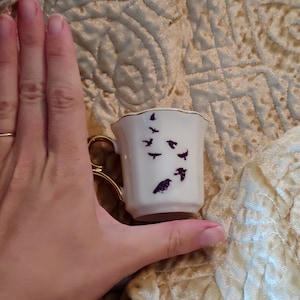 Pode incluir: Uma pequena x&iacute;cara de ch&aacute; branca com uma borda dourada e um desenho de silhueta preta de p&aacute;ssaros voando. A x&iacute;cara est&aacute; sendo segurada em uma m&atilde;o.