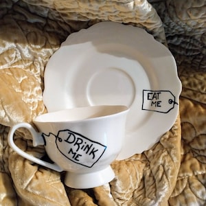 May include: White teacup and saucer with black painted tags that say "Drink Me" and "Eat Me".