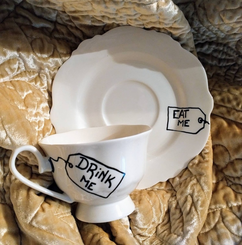 May include: White teacup and saucer with black painted tags that say "Drink Me" and "Eat Me".