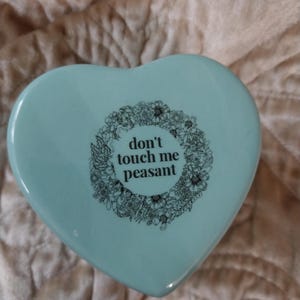 Könnte beinhalten: Hellblaue, herzförmige Schmuckschatulle mit dem Schriftzug "don't touch me peasant" in Schwarz, umgeben von einem floralen Kranz. Die Schatulle liegt auf einer strukturierten, hellbraunen Oberfläche.