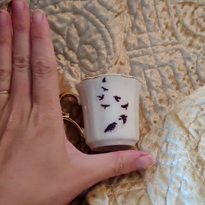 Pode incluir: Uma pequena x&iacute;cara de ch&aacute; branca com uma borda dourada e um desenho de silhueta preta de p&aacute;ssaros voando. A x&iacute;cara est&aacute; sendo segurada em uma m&atilde;o.