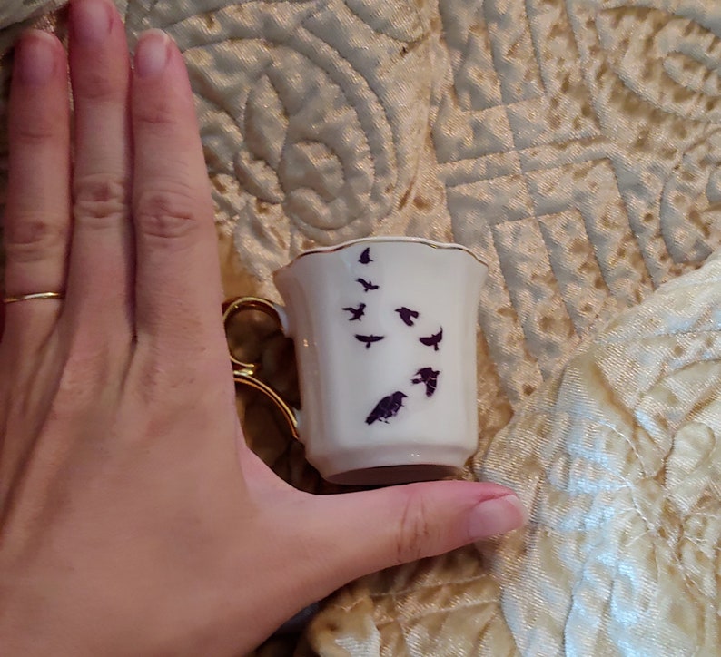 Pode incluir: Uma pequena x&iacute;cara de ch&aacute; branca com uma borda dourada e um desenho de silhueta preta de p&aacute;ssaros voando. A x&iacute;cara est&aacute; sendo segurada em uma m&atilde;o.