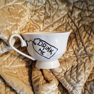 May include: White teacup with a black handle and a black tag that says "Drink Me". The teacup is sitting on a gold fabric background.