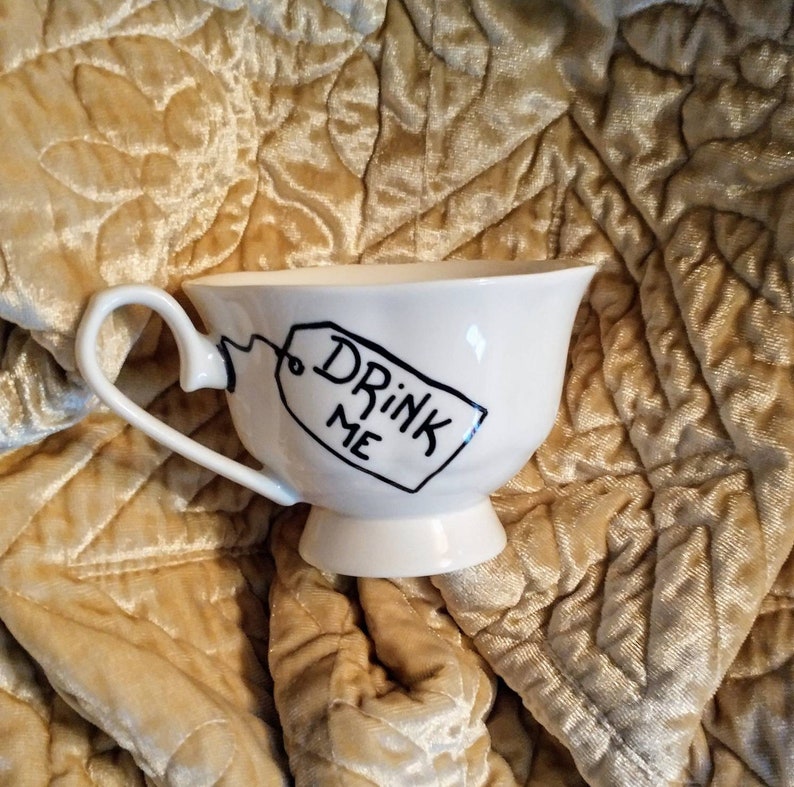 May include: White teacup with a black handle and a black tag that says "Drink Me". The teacup is sitting on a gold fabric background.
