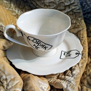May include: White teacup and saucer with black writing. The teacup says "Drink Me" and the saucer says "Eat Me".