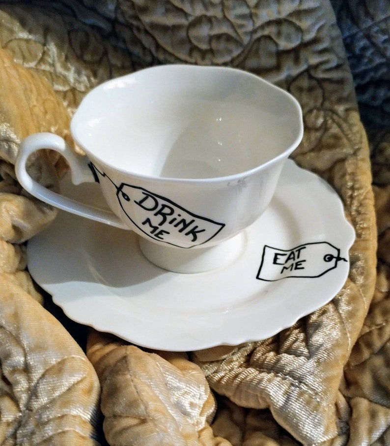 May include: White teacup and saucer with black writing. The teacup says "Drink Me" and the saucer says "Eat Me".