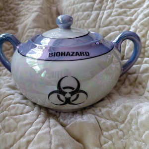 May include: A white ceramic sugar bowl with an iridescent finish, two side handles, and a matching lid. The bowl features a black biohazard symbol on the front and the word BIOHAZARD printed in black capital letters along the upper rim.