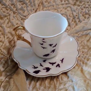 Pode incluir: Um conjunto de ch&aacute;vena e pires branco com aro dourado. A ch&aacute;vena e o pires s&atilde;o decorados com um padr&atilde;o de p&aacute;ssaros roxos em voo.
