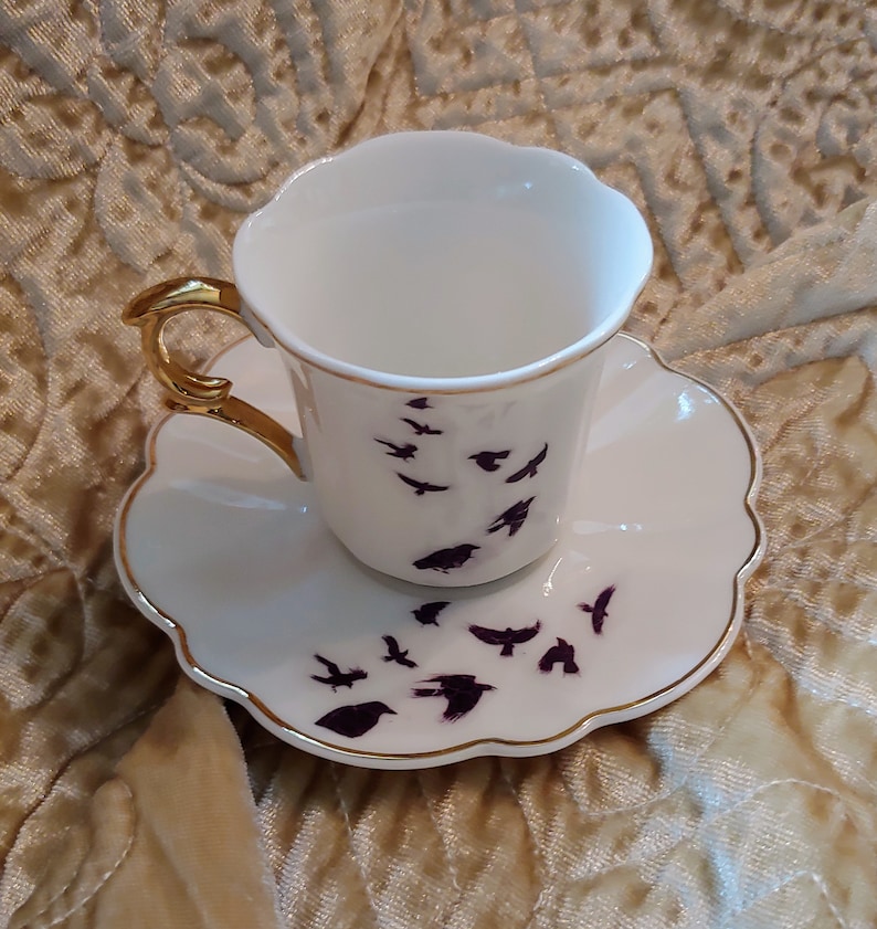 Pode incluir: Um conjunto de ch&aacute;vena e pires branco com aro dourado. A ch&aacute;vena e o pires s&atilde;o decorados com um padr&atilde;o de p&aacute;ssaros roxos em voo.