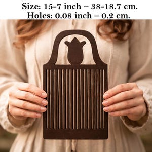 May include: A dark brown wooden weaving tool with a tulip-shaped top and vertical slots. The tool measures 15-7 inches in size and has holes of 0.08 inches. The tool is held in front of a person wearing a cream-colored top.