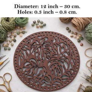 May include: A circular dark brown wooden base with a laser-cut monstera leaf pattern and a perimeter of small holes. Text states: Diameter: 12 inch – 30 cm. Holes: 0.3 inch – 0.8 cm. The base is surrounded by yarn balls, beads, and metal craft tools.