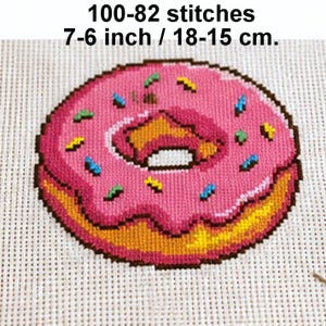 May include: A cross-stitch pattern of a donut with pink frosting and colorful sprinkles on white Aida cloth. The text reads 100-82 stitches and 7-6 inch / 18-15 cm. The donut features a golden-brown base and a dark brown outline.