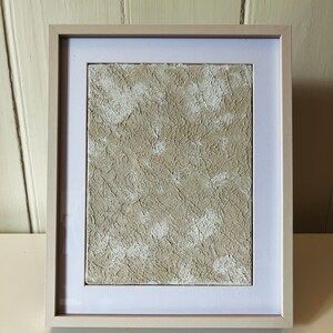 May include: A framed abstract painting with a textured beige and white surface. The painting is in a simple white frame.