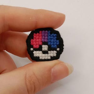 May include: A small circular cross-stitch patch featuring a black border and a segmented design. The top half contains three vertical sections in pink, purple, and blue, while the bottom half is white with a black horizontal line across the center.