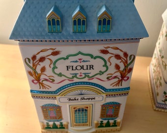 May include: A white ceramic flour canister shaped like a house with a blue roof and a sign that says "Bake Shoppe". The canister has a wheat design and the word "Flour" on the front.