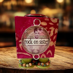 May include: A decorative, handcrafted trinket box with a vibrant fuchsia exterior and a circular pendant. The pendant features a vintage image and the text "rock on sister". The base is adorned with green accents and beaded embellishments.