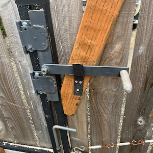 Könnte beinhalten: Nahaufnahme eines schwarzen Metall-Torriegels mit einem Holzgriff. Der Riegel ist an einem Holztor mit einem schwarzen Metallrahmen befestigt. Das Tor besteht aus vertikalen Holzbrettern.