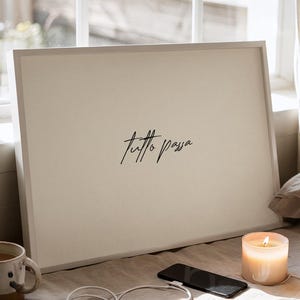 Può includere: Una stampa incorniciata con le parole "Tutto Passa" in corsivo è appoggiata su un letto. Una tazza, un telefono, un cavo di ricarica e una candela accesa sono anch'essi sul letto. La scena è immersa nella luce naturale.