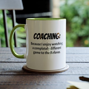 May include: White ceramic coffee mug with a green rim. The mug has black text that reads "COACHING: Because I enjoy watching a completely different game to the Referee."