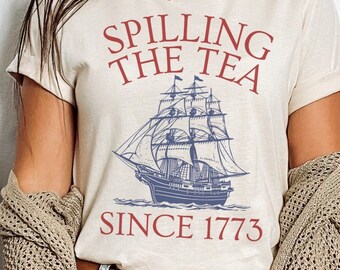 Puede incluir: Una camiseta beige con el texto "Spilling the Tea Since 1773" en rojo y azul. Una ilustración azul y blanca de un barco con velas está en el centro de la camiseta.
