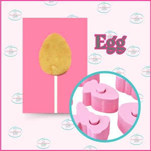 Könnte beinhalten: Ein rosa-weißer Lolli in Form eines Eies. Der Lolli befindet sich auf einem rosa Hintergrund. Es gibt vier rosa Silikonformen in Form von Eiern, Schmetterlingen und Fischen.