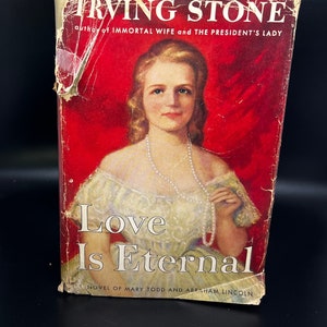 Könnte beinhalten: Ein Vintage-Paperback-Buchcover mit rotem Hintergrund und einem Porträt einer Frau in einem weißen Kleid und einer Perlenkette. Der Titel des Buches lautet "Love Is Eternal" und der Autor ist Irving Stone. Der Untertitel lautet "Roman von Mary Todd und Abraham Lincoln".
