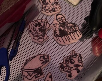 Pode incluir: Uma coleção de seis carimbos de borracha com vários personagens. Os carimbos são rosa e têm tinta preta. Os personagens incluem um fantasma, um homem com uma faca, um peixe, uma personagem de desenho animado e um tubarão.