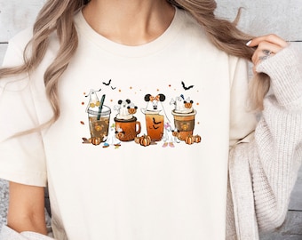 May include: Cream-colored t-shirt featuring a Halloween-themed graphic. The design includes four cartoon coffee cups with ghost and mouse character designs, pumpkins, and falling leaves. The word "Natural" is printed at the top.