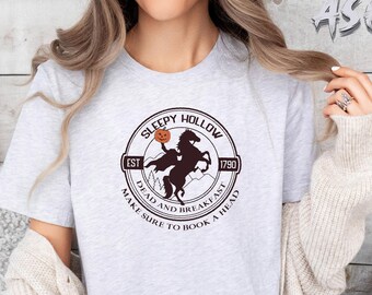 May include: A light gray t-shirt featuring a circular design with the words "Sleepy Hollow" and "Dead and Breakfast." The design includes a horse and rider silhouette, a pumpkin, and the text "Make sure to book a head."