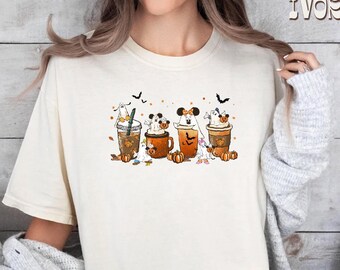 May include: Ivory-colored t-shirt featuring a Halloween-themed graphic. The design showcases four coffee or tea cups with cartoon ghosts and pumpkins. The word "IVORY" is printed in the upper right corner.